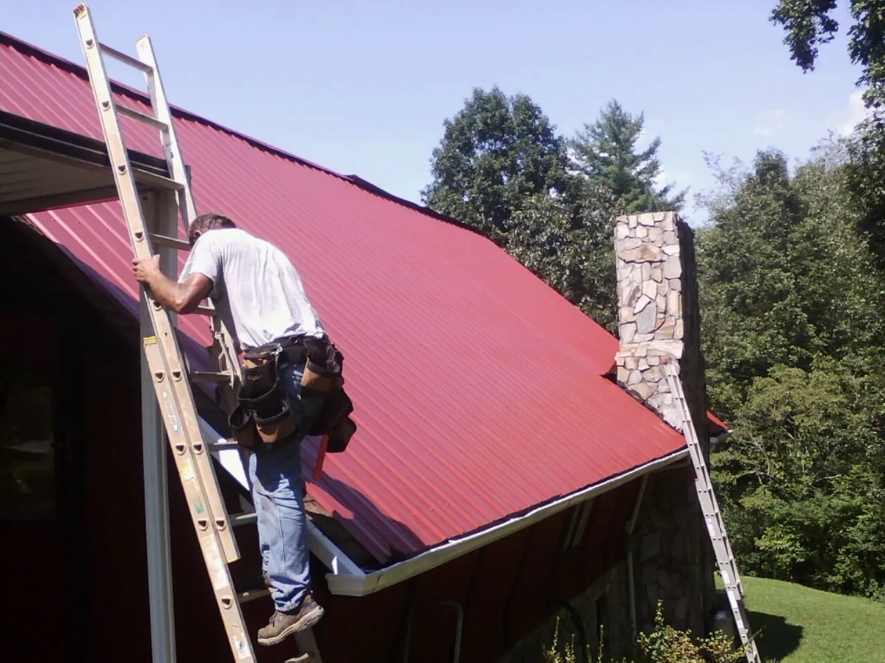 Roofer on a ladder beside a red metal roof and stone chimney — Soffit & Fascia Repair by Roofing Brothers in Leiters Ford